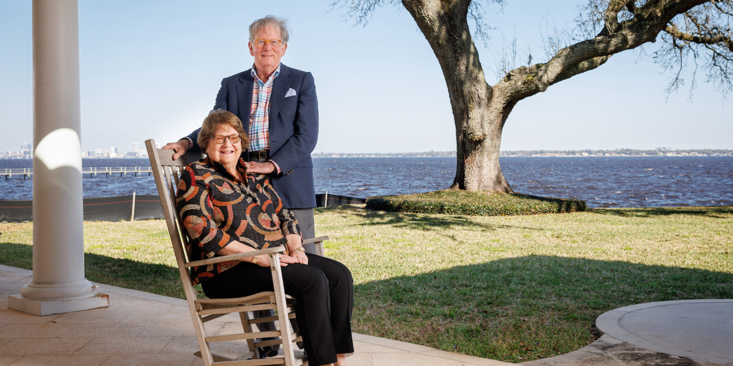 man stands next to wife seated in rocking chair on patio near waterfront