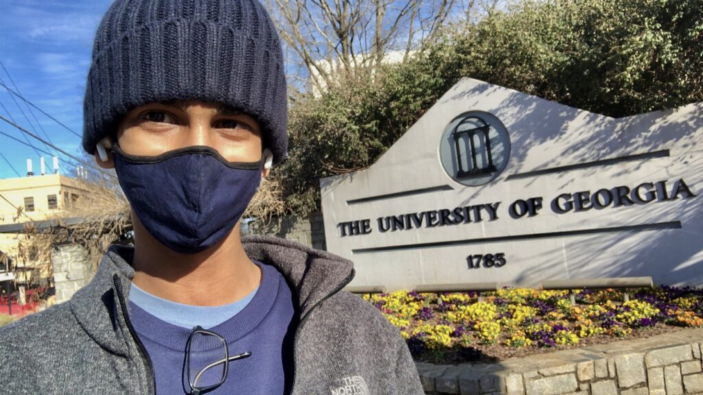 Buhusuru Ranasinghe stands in front a stone UGA sign in a mask and winter hat.